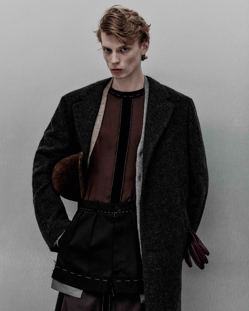 Editorial fashion shot of a young adult male model in a brown semi-sheer shirt, black outwear, wool coat, with a neutral studio backdrop.