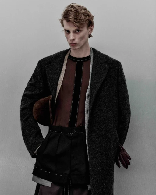 Editorial fashion shot of a young adult male model in a brown semi-sheer shirt, black outwear, wool coat, with a neutral studio backdrop.