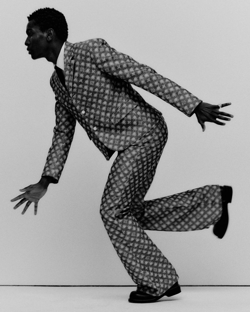 Fashion shot of a Black man in patterned suit, mid-stride against white backdrop, showcasing stylish pose and monochrome aesthetic.