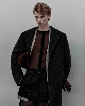 Model Owen Ruppersburg wearing a dark textured overcoat and layered burgundy top against a muted grey studio backdrop.