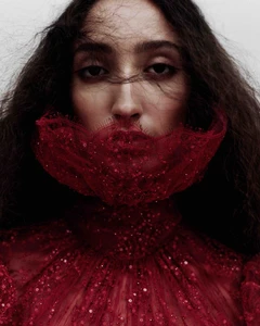 Close-up portrait of a woman with dark wavy hair wearing a structured red lace face covering and garment against a pale background.
