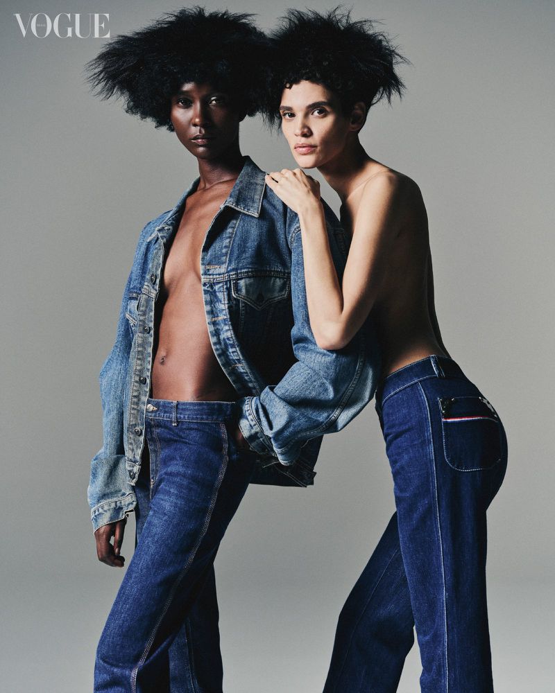 Two fashion models pose in blue jeans and denim jacket in a studio shot for Vogue, showcasing contemporary fashion against a neutral backdrop.