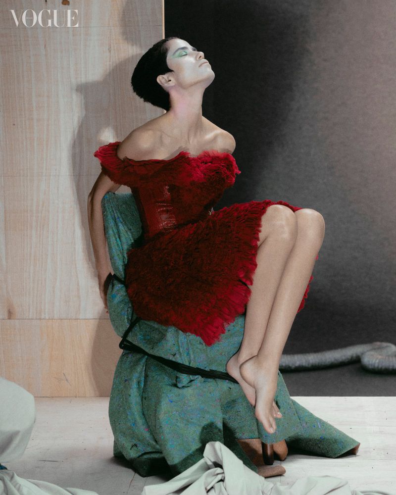 A fashion portrait of a woman in a red dress sitting on a wrapped base in a studio with warm, directional lighting and a Vogue logo visible.