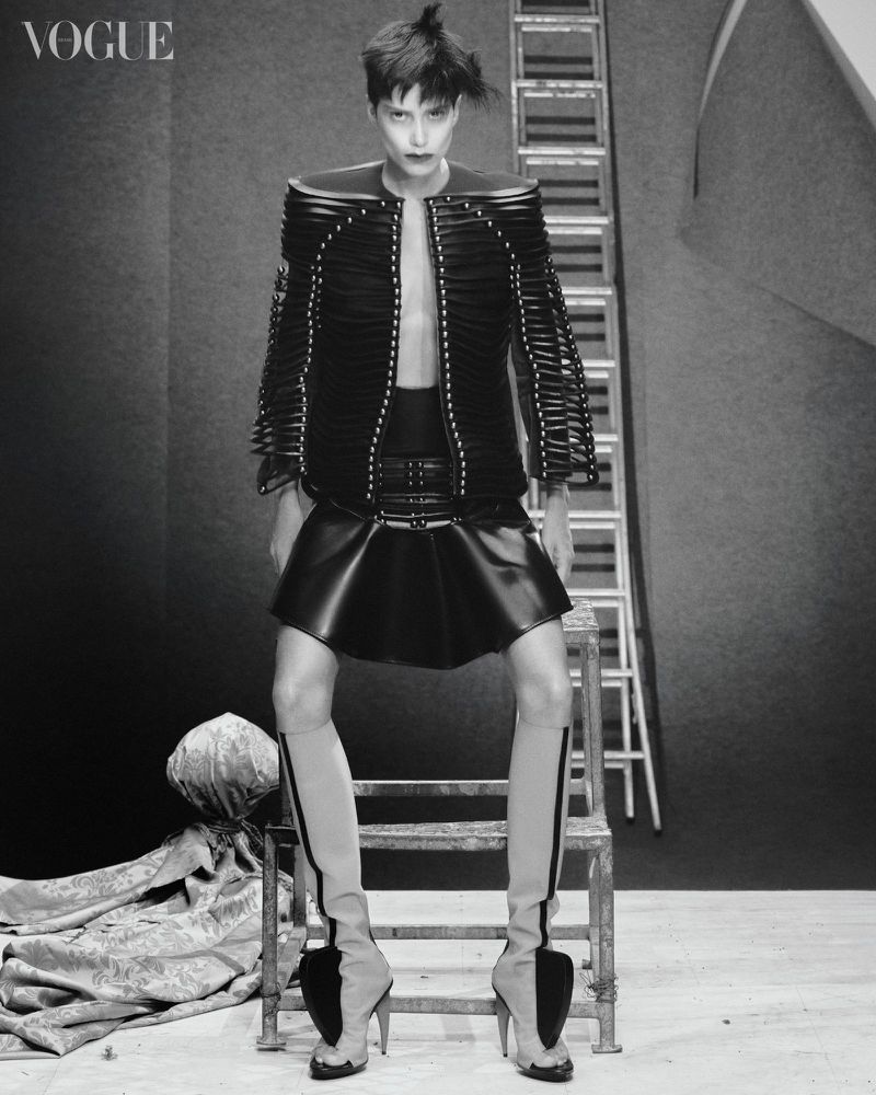 Fashion editorial shot: Woman poses in black jacket, skirt, and sculptural heels on a metal stool with a ladder background, Vogue logo.
