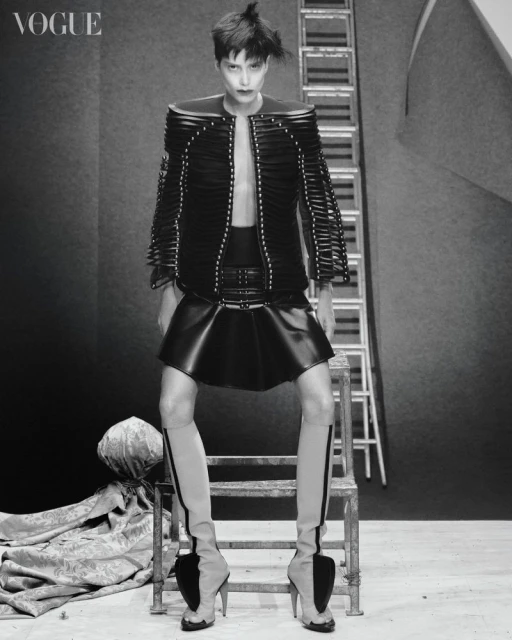 Fashion editorial shot: Woman poses in black jacket, skirt, and sculptural heels on a metal stool with a ladder background, Vogue logo.