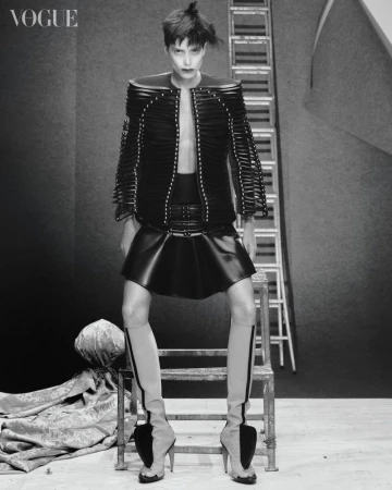 Fashion editorial shot: Woman poses in black jacket, skirt, and sculptural heels on a metal stool with a ladder background, Vogue logo.