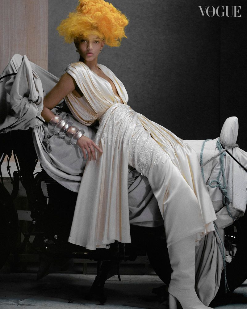 Fashion editorial shot of a model with a voluminous yellow hairpiece, seated in a flowing white gown, against a dark grey backdrop, featuring 'Vogue' logo.