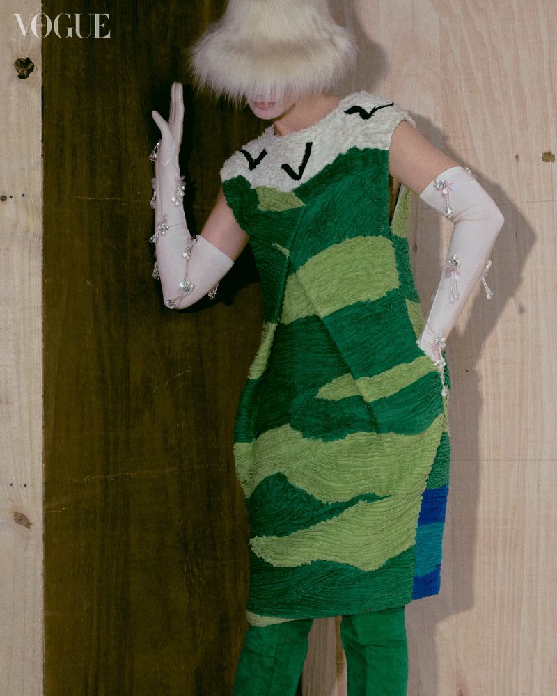 Fashion editorial image featuring a person in a green and cream patterned dress, white gloves, and a furry hat against a wooden background.