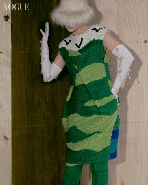 Fashion editorial image featuring a person in a green and cream patterned dress, white gloves, and a furry hat against a wooden background.