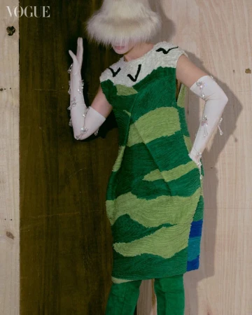 Fashion editorial image featuring a person in a green and cream patterned dress, white gloves, and a furry hat against a wooden background.