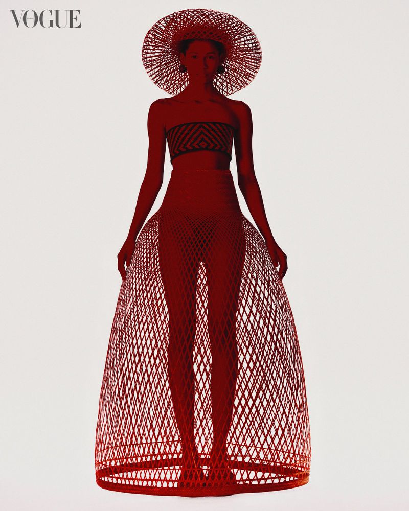 Fashion editorial shot of woman in woven hat, tube top and skirt in front of plain background with Vogue magazine logo in the corner.