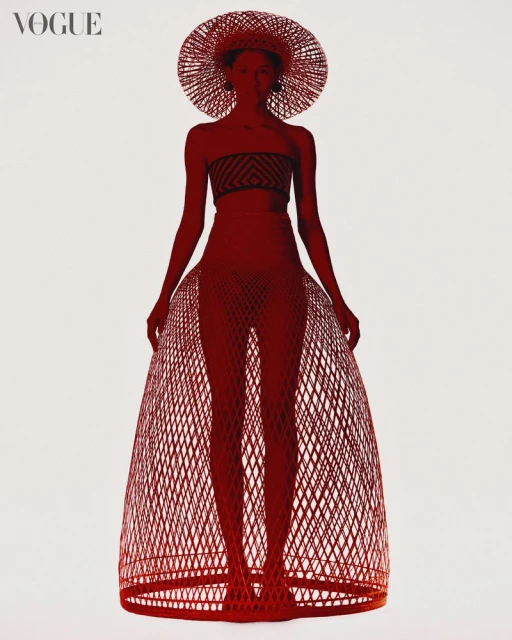 Fashion editorial shot of woman in woven hat, tube top and skirt in front of plain background with Vogue magazine logo in the corner.