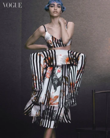 Editorial fashion shot of a blue-haired model in a striped, patterned two-piece outfit in front of a dark studio background with a chair.