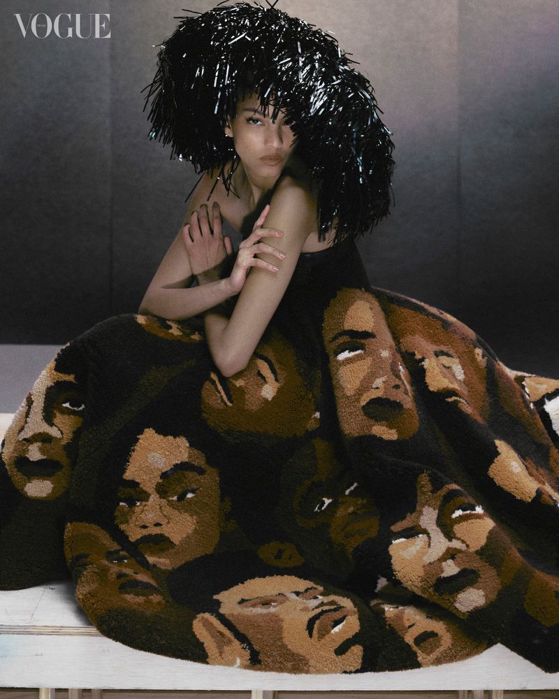 Editorial fashion shot of a person wearing a hat and a dress with faces printed on it, captured in a studio setting, Vogue logo visible.