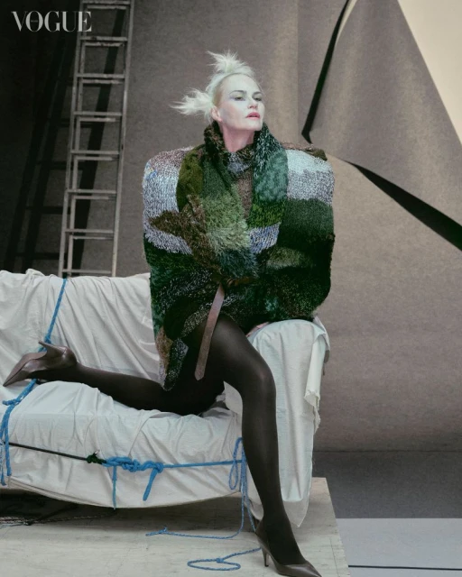 A Vogue fashion shot showcasing a posed woman wearing a unique knit poncho, accessorized with hose and heels, in a styled photo studio.