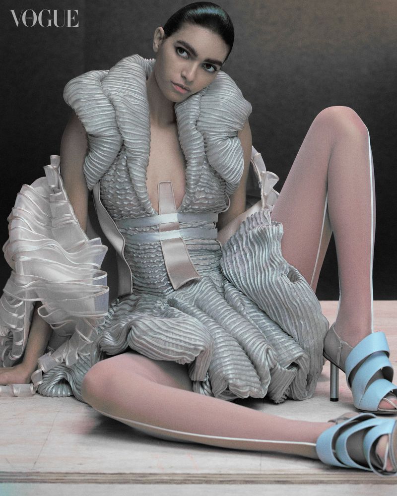 Fashion-forward photo features a model in an avant-garde silver dress with matching high heels, posing in a studio setting for Vogue.