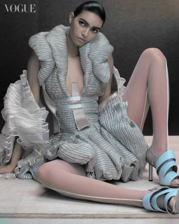 Fashion-forward photo features a model in an avant-garde silver dress with matching high heels, posing in a studio setting for Vogue.