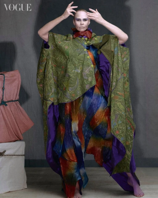 Fashion editorial shot of a woman in a green and multicolored garment striking a pose in front of a gray background with objects to the left.