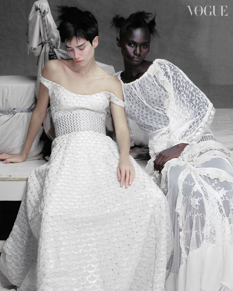Two models in white dresses pose in a fashion editorial with a grey backdrop and Vogue logo visible in the upper-right corner.