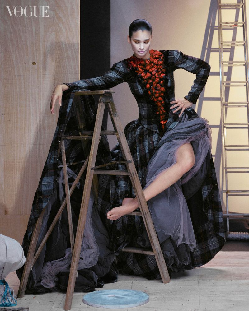 Fashion model in a plaid dress poses on a ladder in a photo studio with soft lighting and a Vogue magazine logo in the upper left.