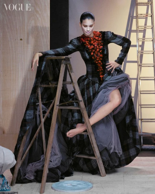 Fashion model in a plaid dress poses on a ladder in a photo studio with soft lighting and a Vogue magazine logo in the upper left.