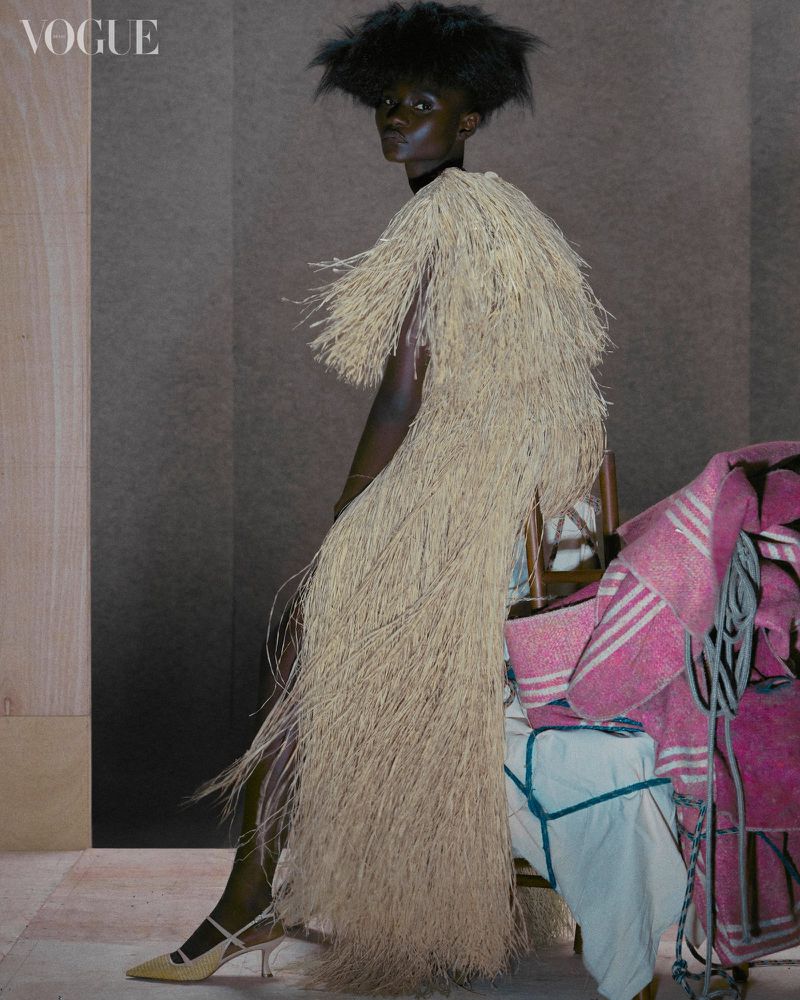 An African-descent fashion model poses in a fringed tan dress for a fashion editorial, striking a pose in an indoor studio setting.