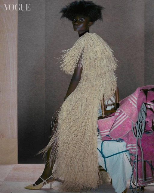 An African-descent fashion model poses in a fringed tan dress for a fashion editorial, striking a pose in an indoor studio setting.