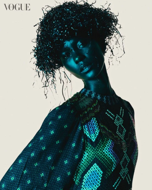 Dark-skinned model with unique hairstyle poses in an editorial fashion shot, wearing patterned sweater against a plain background.