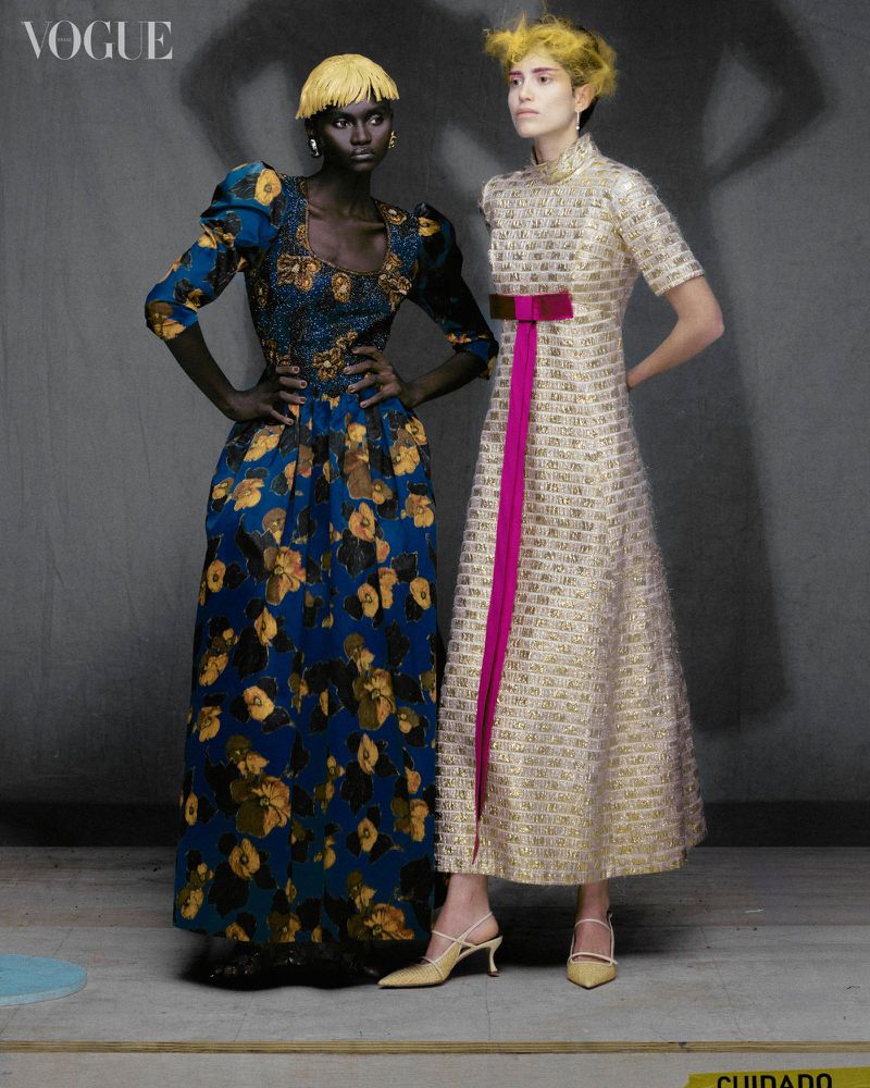Two women in full-length gowns pose in a studio setting; one in a blue floral dress, the other in a gold and pink gown. Vogue logo is visible.
