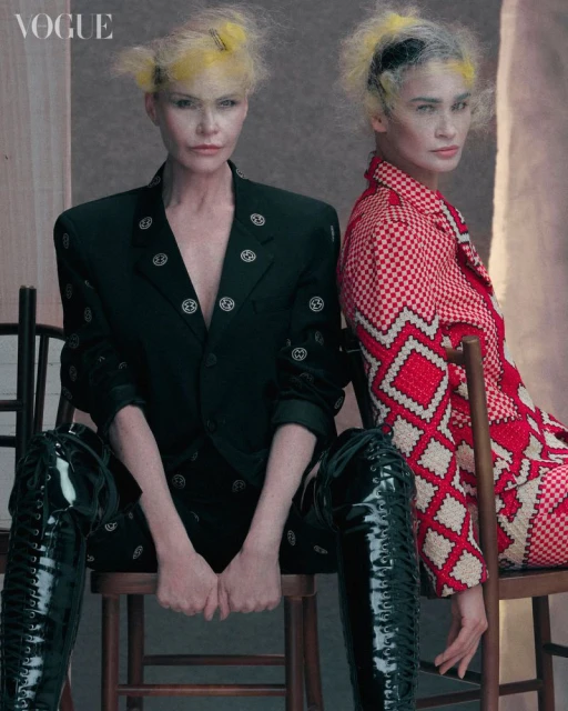 Two fashion models with yellow-tinted hairstyles wearing patterned blazers and shiny thigh-high boots are sitting on wooden chairs.
