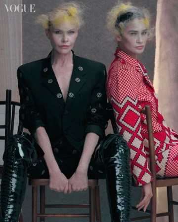 Two fashion models with yellow-tinted hairstyles wearing patterned blazers and shiny thigh-high boots are sitting on wooden chairs.