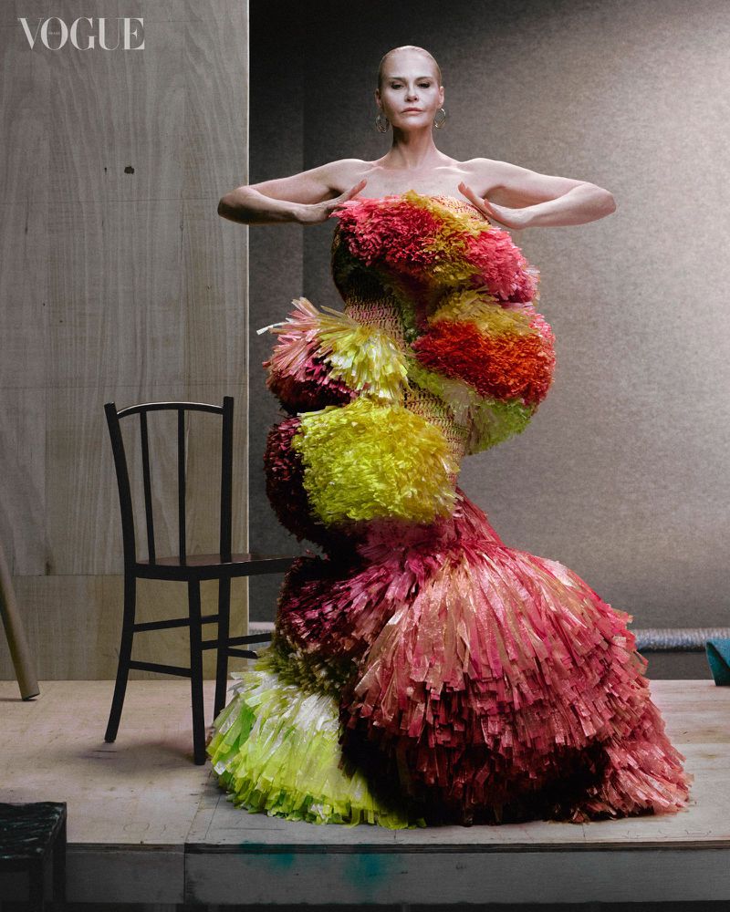 Fashion shot of a model wearing a colorful, frilled dress, posing with raised hands in a studio setting with a chair nearby.