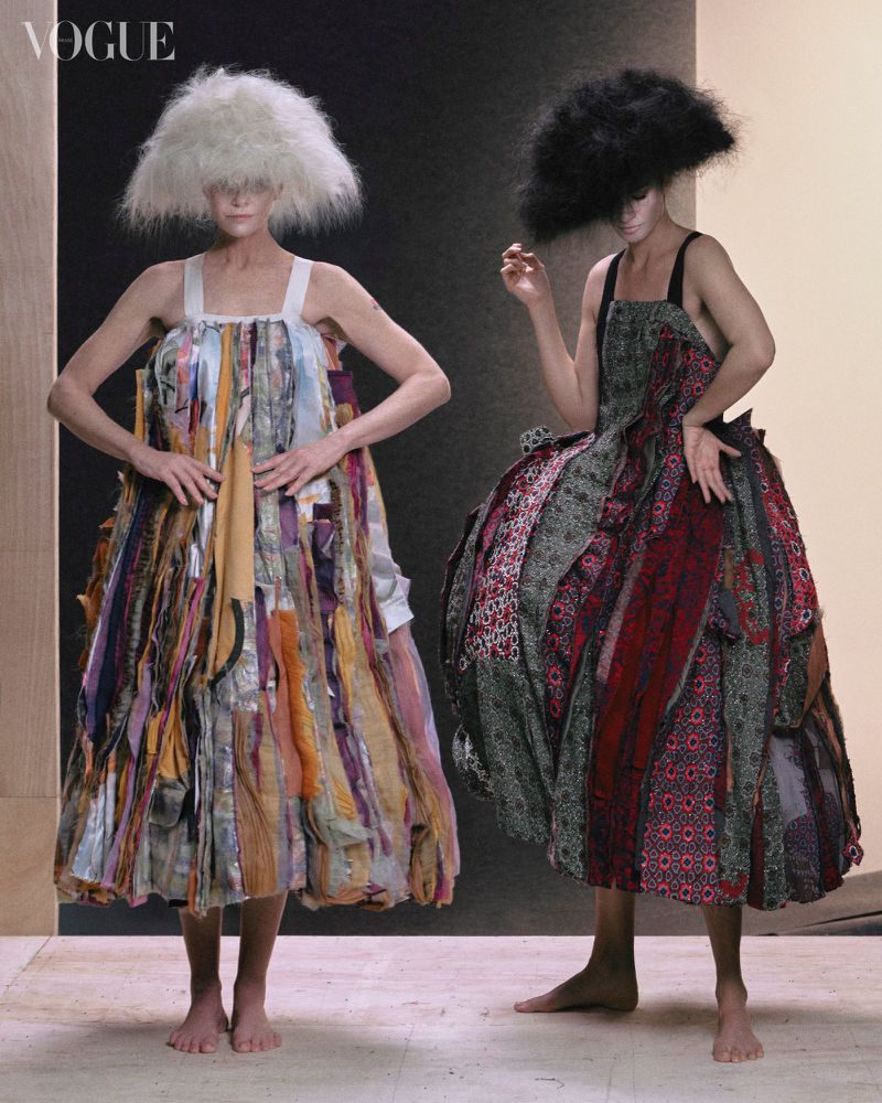 Two fashion models pose indoors wearing large dresses made of colorful fabric strips and big, textured, unusual hairstyles in a studio.