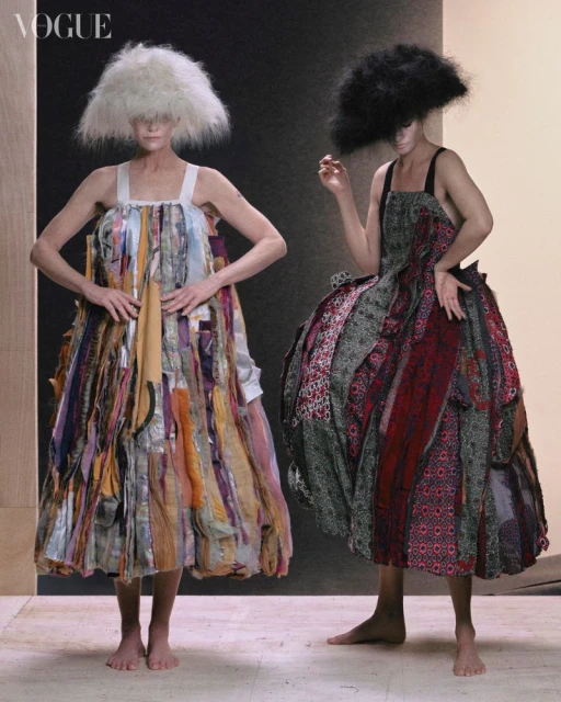 Two fashion models pose indoors wearing large dresses made of colorful fabric strips and big, textured, unusual hairstyles in a studio.