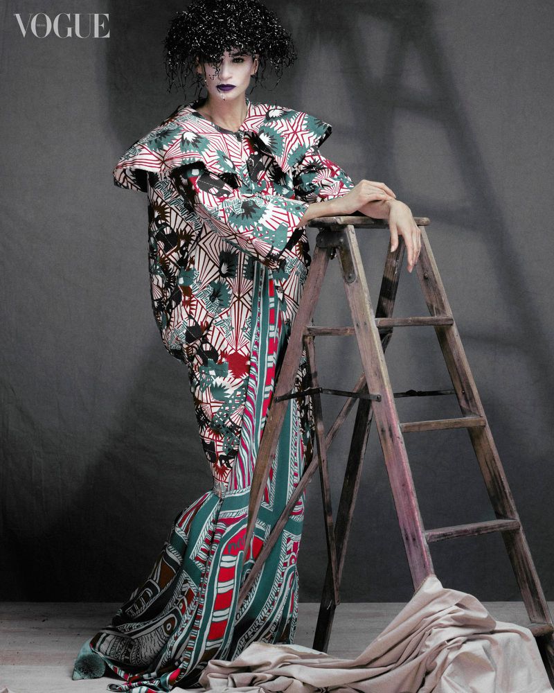 A model in a patterned dress poses next to a ladder in a studio. Fashion editorial photography with dark colors and unique textures.
