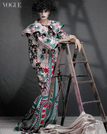A model in a patterned dress poses next to a ladder in a studio. Fashion editorial photography with dark colors and unique textures.