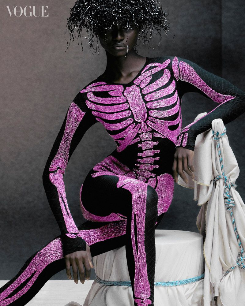 Fashion editorial shot of a Black female model in a black catsuit with a pink skeleton design, sitting indoors in a studio setting.