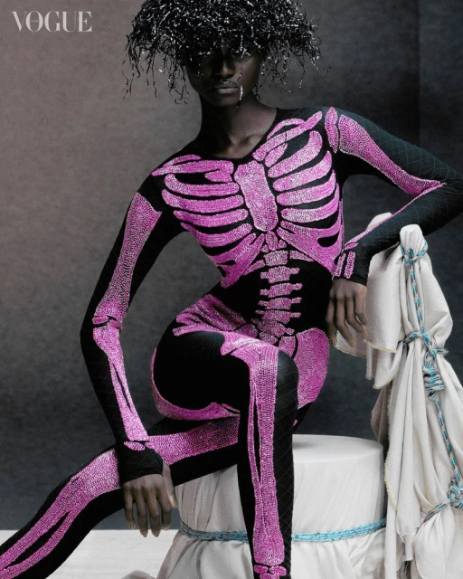 Fashion editorial shot of a Black female model in a black catsuit with a pink skeleton design, sitting indoors in a studio setting.