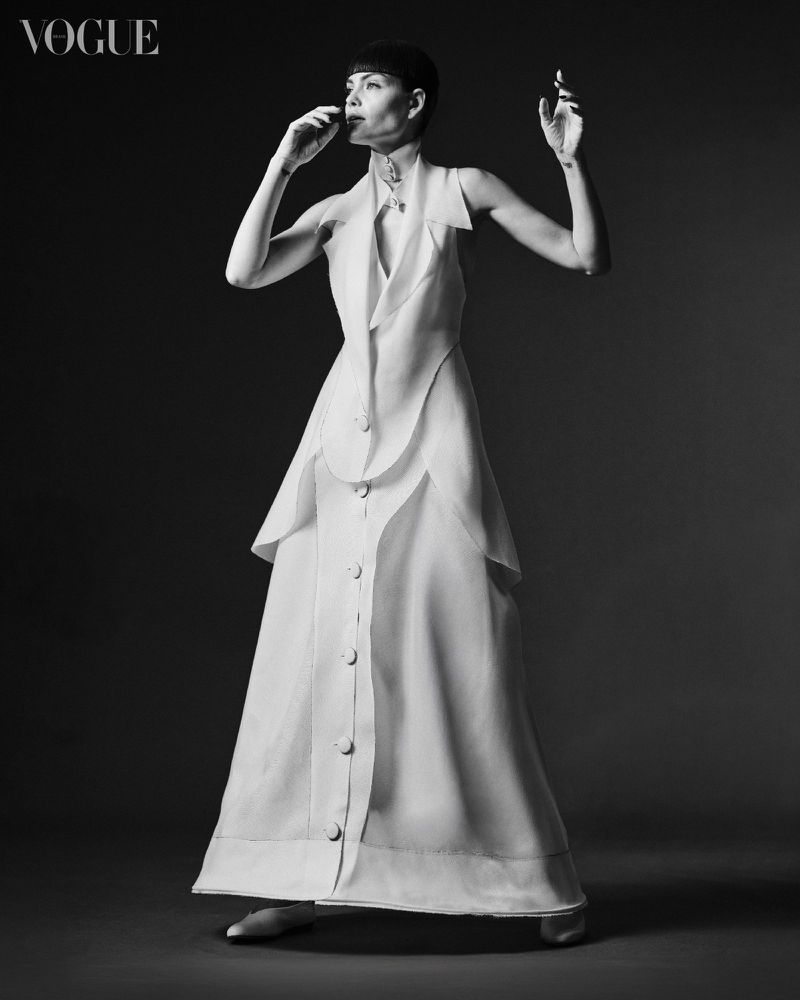 Full-length portrait of a woman in a white dress against a dark background, with Vogue logo in the upper left corner.