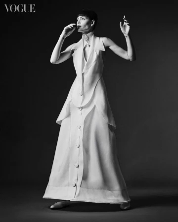 Full-length portrait of a woman in a white dress against a dark background, with Vogue logo in the upper left corner.