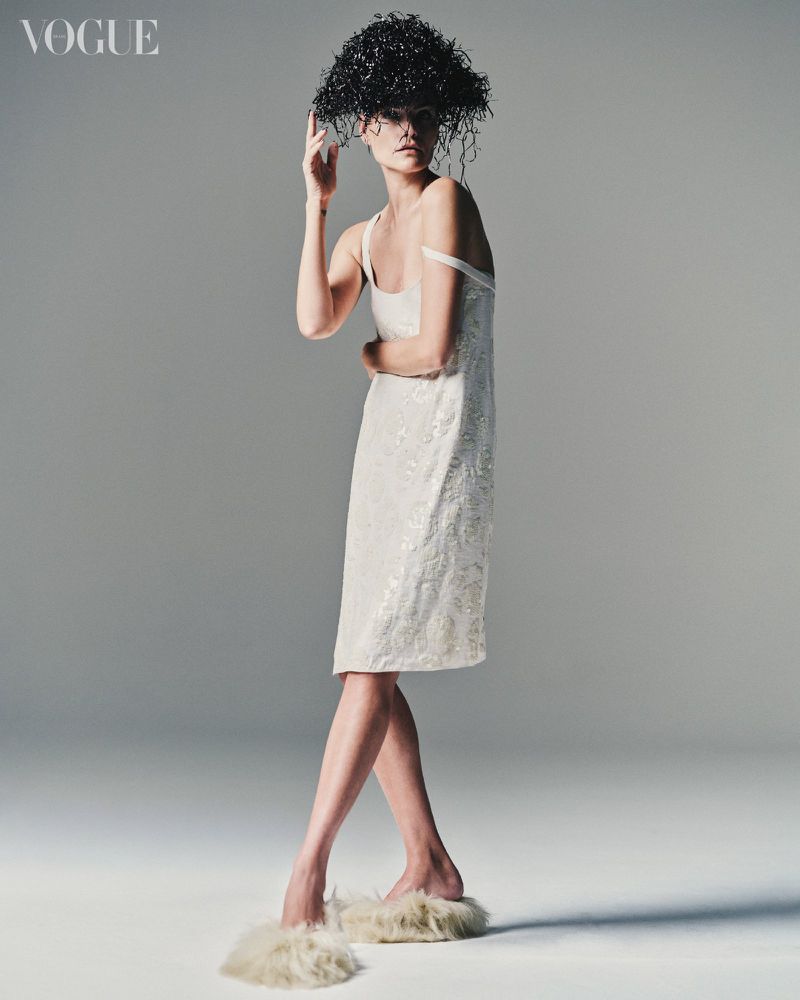Fashion shot of a female model in a sequined dress and unique headpiece, posing in a photo studio with a neutral gray background and soft lighting.