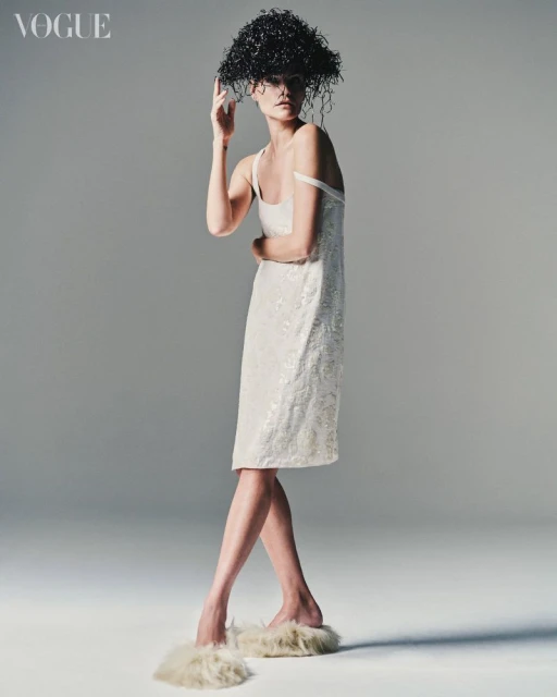 Fashion shot of a female model in a sequined dress and unique headpiece, posing in a photo studio with a neutral gray background and soft lighting.