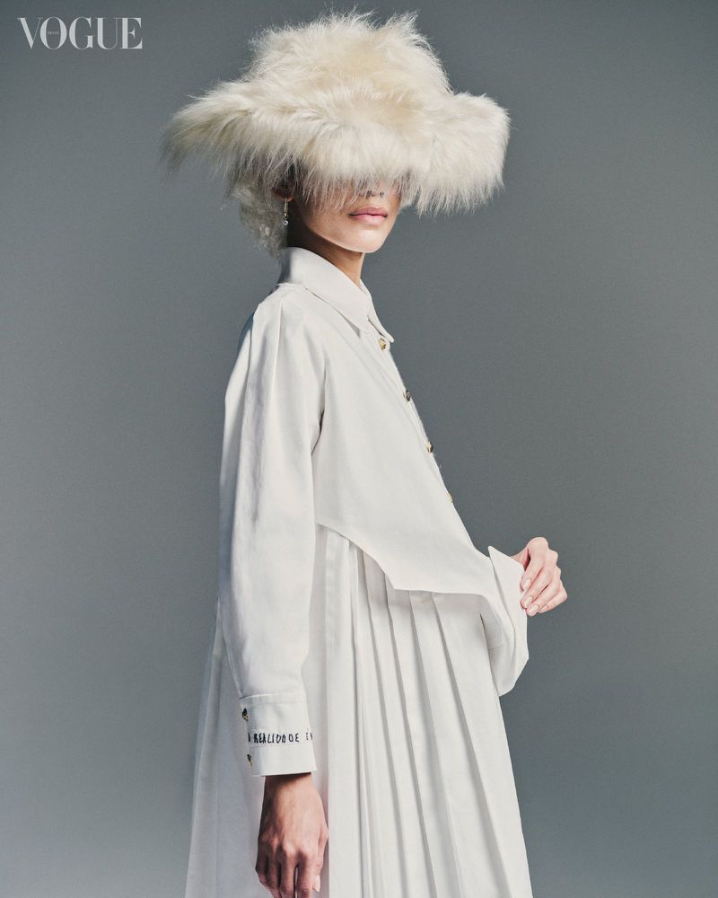 Model wearing an oversized fluffy white fur hat obscuring her eyes and a pleated white shirt dress against a grey studio backdrop.