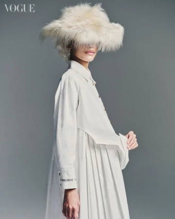 Model wearing an oversized fluffy white fur hat obscuring her eyes and a pleated white shirt dress against a grey studio backdrop.