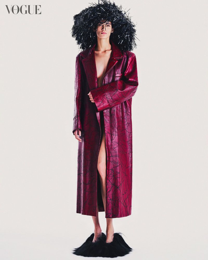 Fashion shot of a slender figure in a long red coat, adorned with a black headpiece and fuzzy slippers, against a neutral backdrop.