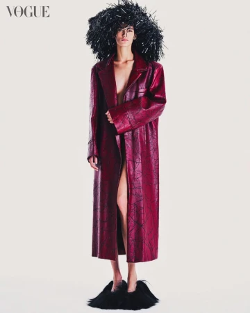 Fashion shot of a slender figure in a long red coat, adorned with a black headpiece and fuzzy slippers, against a neutral backdrop.