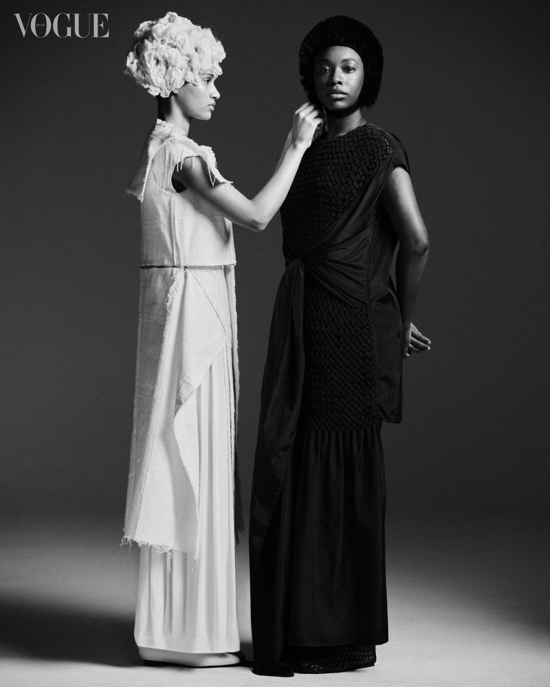 Two fashion models in contrasting black and white dresses pose in a studio. One adjusts the other's collar in a Vogue editorial shot.