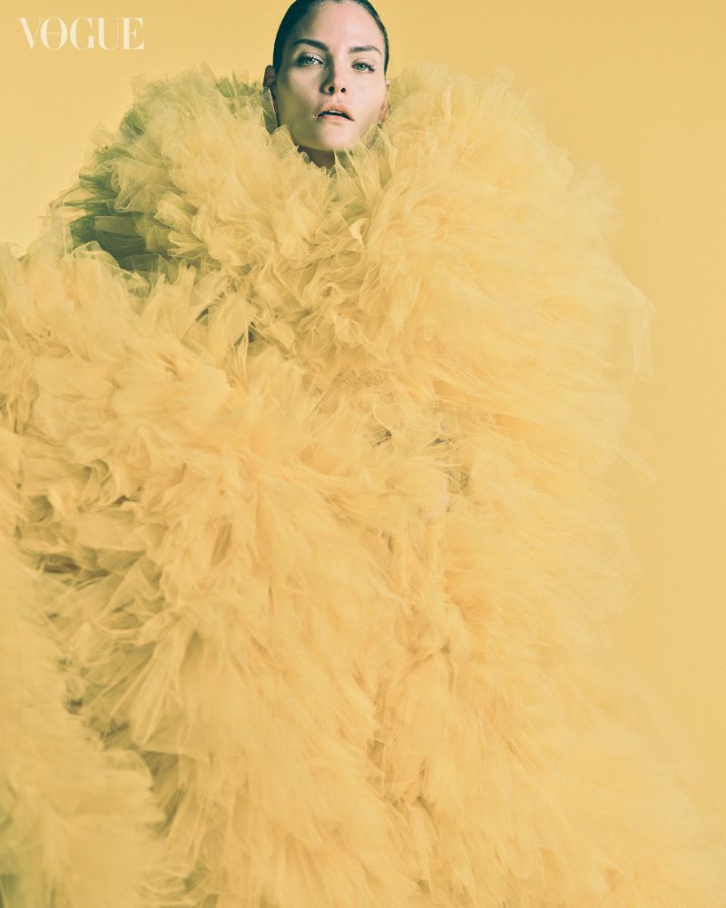 Fashion editorial shot of an adult woman in a large, frilly, yellow tulle dress against a matching yellow background in the photo studio.