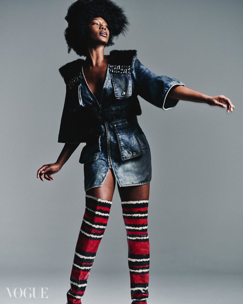Fashion editorial shot of a Black woman in denim dress, fur accents, and thigh-high striped boots posing against a neutral gray background.