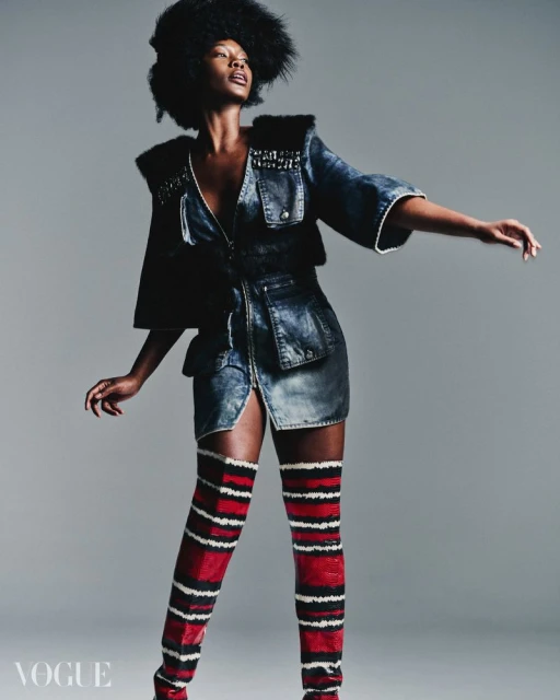 Fashion editorial shot of a Black woman in denim dress, fur accents, and thigh-high striped boots posing against a neutral gray background.